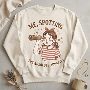 May include: Cream-colored sweatshirt with a graphic of a cartoon girl looking through a telescope. The text reads "ME, SPOTTING THE ABSOLUTE AUDACITY". The girl wears a red headband and a striped shirt. Hearts and stars surround the text.