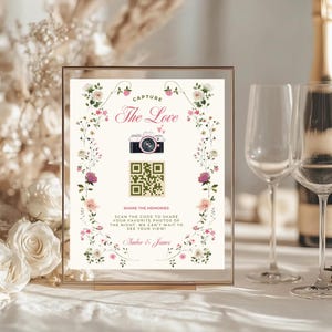 Könnte beinhalten: Ein gerahmtes Hochzeitsschild mit floralen Verzierungen und dem Text "Capture The Love". Das Schild zeigt eine Kamera-Illustration, einen QR-Code und die Namen "Amber & James". Es steht auf einem Tisch mit Sektgläsern.