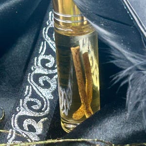 May include: A clear glass roll-on bottle with a gold cap, filled with a light amber liquid and two cinnamon sticks. The bottle is set against a dark background with black feathers and a decorative ribbon.