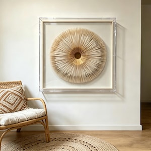 May include: A framed, circular wall art piece with a layered, textured design in shades of beige and gold. The artwork is encased in a clear acrylic frame and mounted on a white wall. A woven chair and rug are in the foreground.