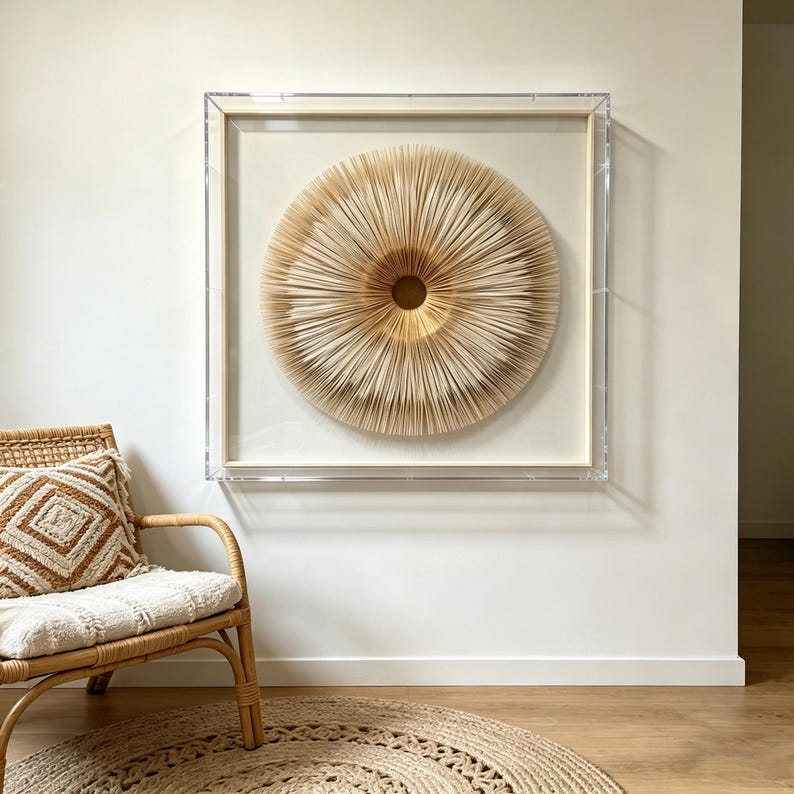 May include: A framed, circular wall art piece with a layered, textured design in shades of beige and gold. The artwork is encased in a clear acrylic frame and mounted on a white wall. A woven chair and rug are in the foreground.
