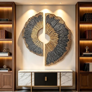 May include: A two-piece circular wall sculpture featuring a textured black and gold sunburst pattern. The artwork is mounted on a wall above a white marble console table with black cabinet doors, flanked by two tall wooden bookshelves with lighting.