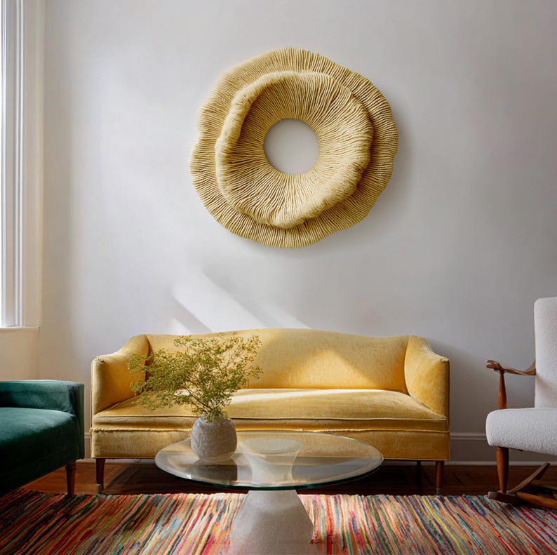 May include: A large, circular wall art piece with a textured, layered design in a light beige color. The artwork hangs on a white wall above a yellow sofa. The room also features a glass coffee table and a colorful rug.