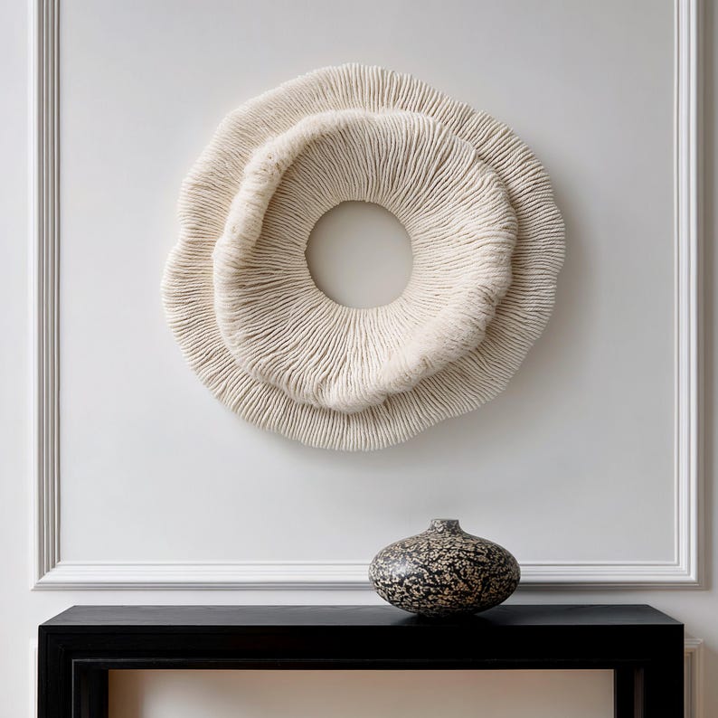 May include: A cream-colored, circular wall art piece with a textured, layered design, resembling a coral formation. Below, a small, speckled vase sits on a black table. The wall is white with decorative molding.