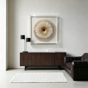 May include: A modern living room setup featuring a dark brown wooden cabinet with vertical grooves, a black lamp, and a framed circular art piece in neutral tones. A white rug and a brown leather sofa complete the scene.