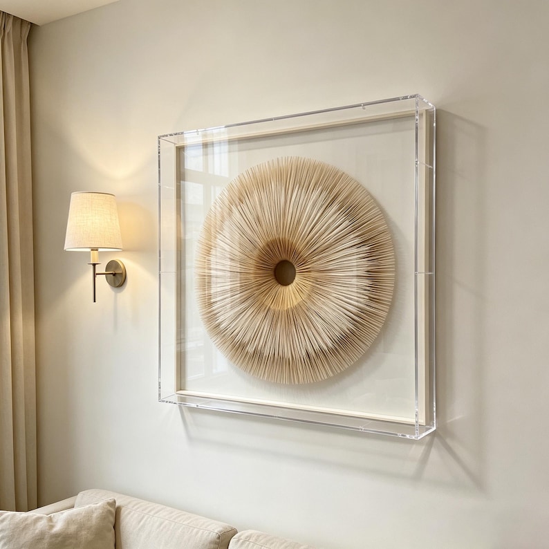 May include: A framed, circular wall art piece with a layered, textured design in shades of beige and tan. The artwork is encased in a clear acrylic frame, creating a modern and minimalist aesthetic. The piece is mounted on a neutral-colored wall.