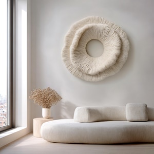 May include: A cream-colored, circular wall art piece with a textured, layered design resembling coral. The art hangs above a cream-colored sofa with a matching cylindrical pillow and a square pillow. A small wooden side table holds a vase with dried flowers.