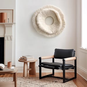 May include: A modern living room scene featuring a large, cream-colored, circular wall art piece. A black leather chair with wooden armrests sits on a patterned rug. A small wooden side table holds books, and a fireplace is visible.