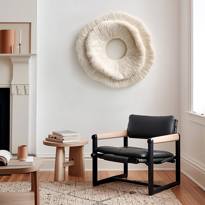 May include: A modern living room scene featuring a large, cream-colored, circular wall art piece. A black leather chair with wooden armrests sits on a patterned rug. A small wooden side table holds books, and a fireplace is visible.