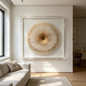 May include: A large, circular wall art piece with a layered, radial design in shades of beige and gold. The artwork is encased in a clear, rectangular frame and mounted on a white wall. The room features a neutral color palette with a sofa and rug.