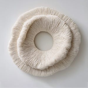 May include: A cream-colored, circular wall art piece with a textured, ridged surface. The design features two overlapping rings, creating a layered effect with a central opening. The piece has a natural, organic aesthetic.