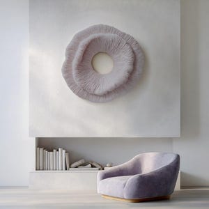 May include: A light purple, circular wall art piece with a textured, layered design hangs above a matching armchair. The artwork has a central opening and is mounted on a white wall. A built-in shelf below holds books and decorative items.