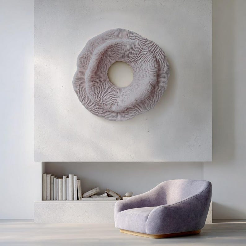 May include: A light purple, circular wall art piece with a textured, layered design hangs above a matching armchair. The artwork has a central opening and is mounted on a white wall. A built-in shelf below holds books and decorative items.