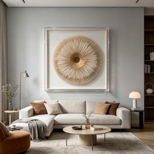 May include: A modern living room with a light gray wall and a large, framed circular art piece in shades of beige and tan. A cream-colored sectional sofa is adorned with brown and beige pillows. A round coffee table and a side table with a lamp complete the scene.