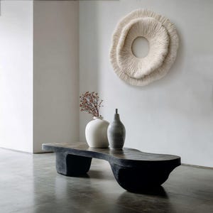 May include: A modern living room scene featuring a black, sculptural coffee table with a dark, textured top. Two ceramic vases and a dried floral arrangement sit on the table. A large, circular, textured wall hanging is mounted on the white wall.