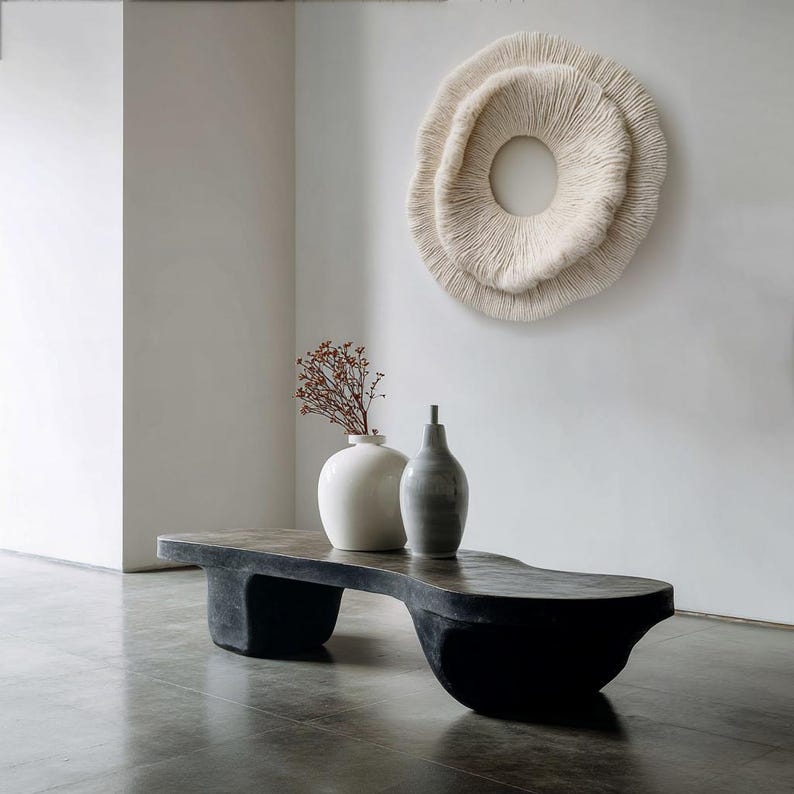 May include: A modern living room scene featuring a black, sculptural coffee table with a dark, textured top. Two ceramic vases and a dried floral arrangement sit on the table. A large, circular, textured wall hanging is mounted on the white wall.