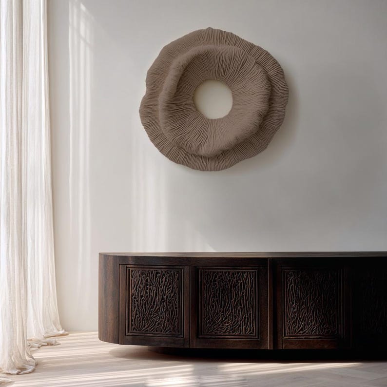 May include: A large, circular, textured wall art piece in a neutral taupe color, mounted on a white wall. Below, a dark wood cabinet with carved, organic patterns on the doors. The cabinet has a curved shape.