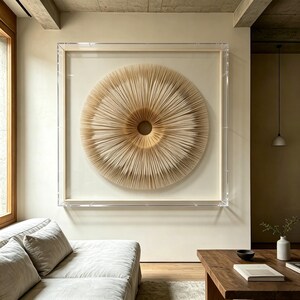 May include: A large, circular wall art piece with a layered, radial design. The artwork is framed in a clear acrylic box, showcasing a textured, beige and tan color palette. The piece hangs above a light-colored sofa and wooden coffee table.