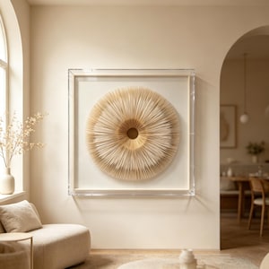 May include: A framed, circular wall art piece with a layered, radial design. The artwork features a light beige color palette with a central, golden-hued core. The piece is encased in a clear, rectangular frame, creating a modern aesthetic.