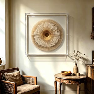 May include: A framed, circular wall art piece with a layered, textured design in shades of beige and gold. The artwork is encased in a clear acrylic frame, creating a modern and elegant aesthetic. The piece is displayed in a room with a vintage chair and table.