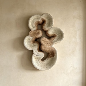 May include: Abstract wall art sculpture in neutral tones. The piece features a layered design with a flowing, organic shape. The sculpture is primarily cream and light brown, creating a textured, three-dimensional effect. It is mounted on a beige wall.