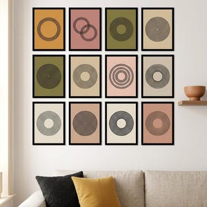 May include: A gallery wall arrangement of twelve rectangular framed prints featuring black line art of concentric circles and geometric patterns on solid backgrounds of mustard yellow, muted pink, olive green, and beige, displayed in a grid.