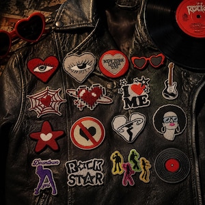 Può includere: Una giacca in pelle nera decorata con varie toppe ricamate, tra cui cuori con occhi, una ragnatela, un cuore pixelato, un cerchio New York Loves You, una chitarra, una persona con afro e scritte come I Love Me, Rock Star e Downtown Girl.