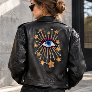 K&ouml;nnte beinhalten: Eine schwarze Lederjacke mit einem gro&szlig;en, gestickten Aufn&auml;her auf der R&uuml;ckseite. Der Aufn&auml;her zeigt ein zentrales blau-wei&szlig;es Augenmotiv, umgeben von strahlenden goldenen Paillettensternen und kleinen silbernen Kreuzakzenten als Grafik.