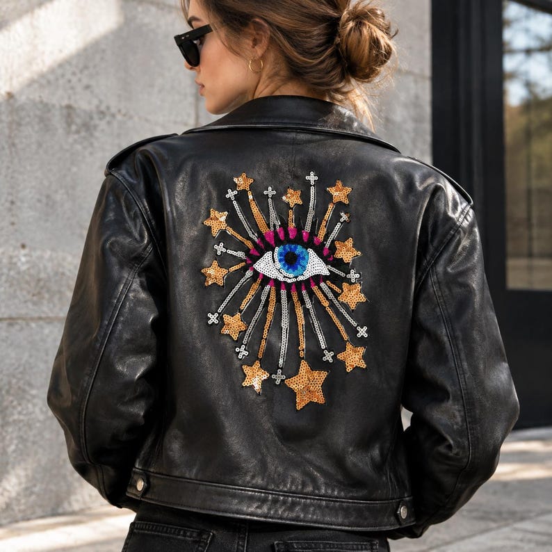 K&ouml;nnte beinhalten: Eine schwarze Lederjacke mit einem gro&szlig;en, gestickten Aufn&auml;her auf der R&uuml;ckseite. Der Aufn&auml;her zeigt ein zentrales blau-wei&szlig;es Augenmotiv, umgeben von strahlenden goldenen Paillettensternen und kleinen silbernen Kreuzakzenten als Grafik.