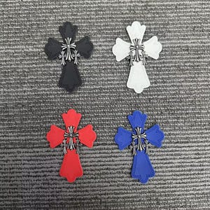 May include: Four cross-shaped patches in black, white, red, and blue. Each patch features a decorative stitched border and a central metallic silver-toned emblem consisting of three small crosses arranged in a vertical cluster at the center.