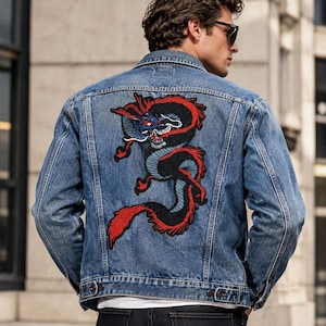 May include: A medium-wash blue denim jacket featuring a large embroidered patch on the back. The patch depicts a stylized black, blue, and red dragon with intricate detailing, positioned centrally across the back panel of the garment.