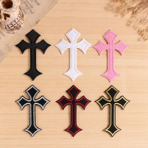 Può includere: Un set di sei toppe ricamate a forma di croce disposte su due file. La fila superiore presenta croci a tinta unita nere, bianche e rosa. La fila inferiore include croci nere con bordi e contorni ricamati in argento, rosso e oro.