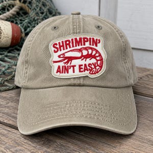 Puede incluir: Una gorra de béisbol color caqui con un parche bordado blanco que muestra un gráfico de un camarón rojo y el texto SHRUMPIN' AIN'T EASY. La gorra tiene una visera curva, ojales cosidos en la corona y un panel frontal estructurado de tela desgastada.