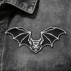 Puede incluir: Parche de murciélago negro con detalles bordados en blanco, que muestra una cara de murciélago y alas extendidas. El parche está cosido en una chaqueta vaquera gris, creando un tema gótico o de Halloween.