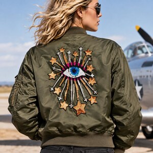 K&ouml;nnte beinhalten: Eine olivgr&uuml;ne Bomberjacke mit einem gro&szlig;en, gestickten Aufn&auml;her auf der R&uuml;ckseite. Das Design zeigt ein zentrales blaues Auge, umgeben von strahlenden, goldfarbenen Paillettensternen und wei&szlig;en Linien, die einen Sonneneffekt mit rosa Akzenten erzeugen.