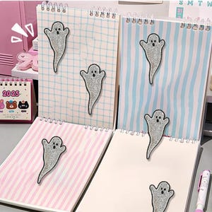 Op de afbeelding: Vier spiraalgebonden notitieboekjes met spookvormige versieringen. De notitieboekjes hebben verschillende pagina patronen: geruit, gestreept en blanco. De spookontwerpen zijn zilverkleurig met zwarte stipogen en een mond. Een kleine kalender is zichtbaar.