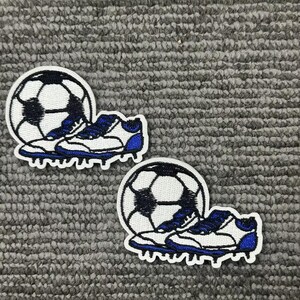 May include: Two embroidered patches depicting a football and a football boot. The football is white with black pentagons. The boot is white with blue detailing. The patches are on a grey textured background.