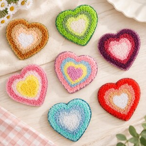 Puede incluir: Siete posavasos con forma de corazón y patrones de colores concéntricos. El conjunto incluye corazones en tonos marrón, verde lima, morado, rosa, amarillo, azul claro y rojo, cada uno con una superficie texturizada de bucle y bordes brillantes.