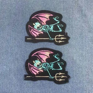 May include: Two embroidered patches featuring a skeleton with teal bones, a teal skull, and pink wings. The skeleton is holding a trident. The patches are on a black background.