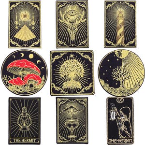 Puede incluir: Una colección de nueve parches bordados sobre tela negra con diseños en hilo dorado, incluyendo una pirámide, un ojo, un faro, setas rojas, un árbol, sol y luna, una linterna, un cáliz y una figura encapuchada con el texto The Hermit.