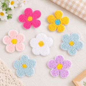 以下が含まれることがあります： 質感のあるふわふわした表面を持つ、7つの花形のワッペン。鮮やかなピンク、青い中心を持つ黄色、淡いピンク、白、水色、紫など、さまざまな色があり、それぞれ対照的な色の中心部が特徴です。