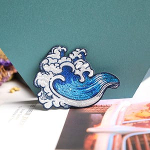 May include: Embroidered patch featuring a stylized ocean wave design. The wave is depicted in shades of blue and white, with intricate stitching details. The patch is likely for decorative purposes, suitable for clothing or accessories.