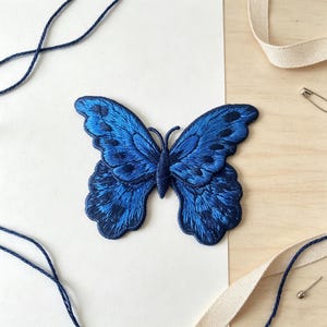 May include: An embroidered butterfly patch featuring shades of deep navy and royal blue thread. The design displays intricate wing patterns with dark blue accents and a central body, showcasing detailed stitching throughout the entire insect shape.