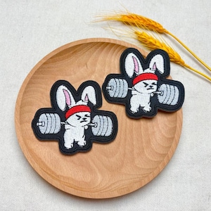 May include: Two embroidered patches showing a white bunny wearing a red headband, lifting a barbell. The patches have a black border and are set on a wooden plate. The barbell has silver weights. The bunny patches are a fun, sporty design.