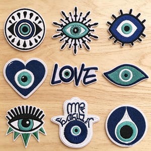 May include: A collection of nine embroidered patches featuring various eye motifs, a heart shape, and the word LOVE. The designs utilize shades of blue, teal, black, and white. Each patch has a distinct shape, including circles, stars, and a hamsa hand.