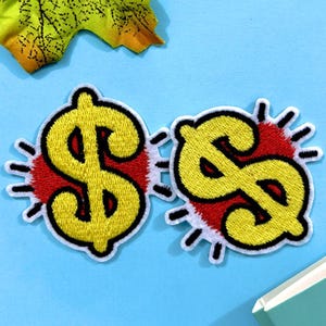 May include: Two embroidered patches featuring a yellow dollar sign ($) with a black outline, set against a red burst design. The patches are on a light blue surface, suggesting a theme of finance or wealth.