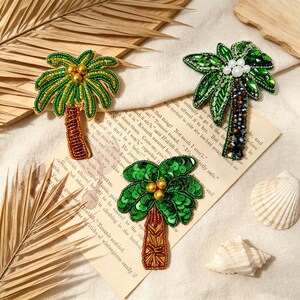 Puede incluir: Tres broches de palmera con intrincados abalorios y lentejuelas. Un broche tiene frondas de cuentas verdes y tronco marrón, otro tiene hojas de cristal verde con perlas blancas, y el tercero tiene frondas de lentejuelas verdes y tronco dorado.