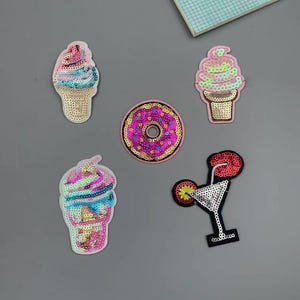 May include: A collection of sequined patches featuring ice cream cones, a donut, and a cocktail. The patches are in various colors, including pink, gold, and blue. The patches are on a gray background.