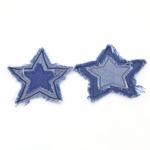 May include: Two denim star patches with frayed edges. Each star has a layered design with a lighter blue inner star and a darker blue outer star. Black stitching outlines the inner stars.