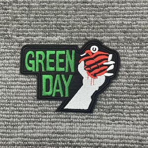 以下が含まれることがあります： 黒い背景に緑色のブロック文字で「GREEN DAY」と書かれた刺繍パッチ。パッチには、血が滴る赤と黒の手榴弾を握る白い手も描かれています。